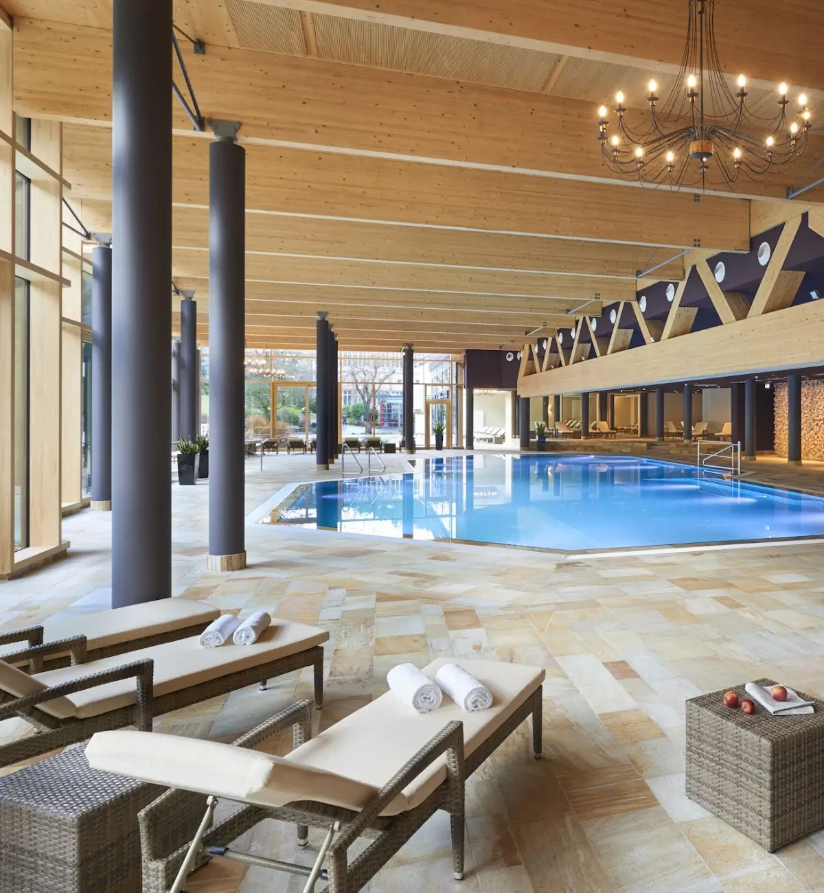 Elegant indoor pool area at Hotel Therme Bad Teinach, featuring a modern yet warm design with wooden beams, floor-to-ceiling windows, and a grand chandelier.