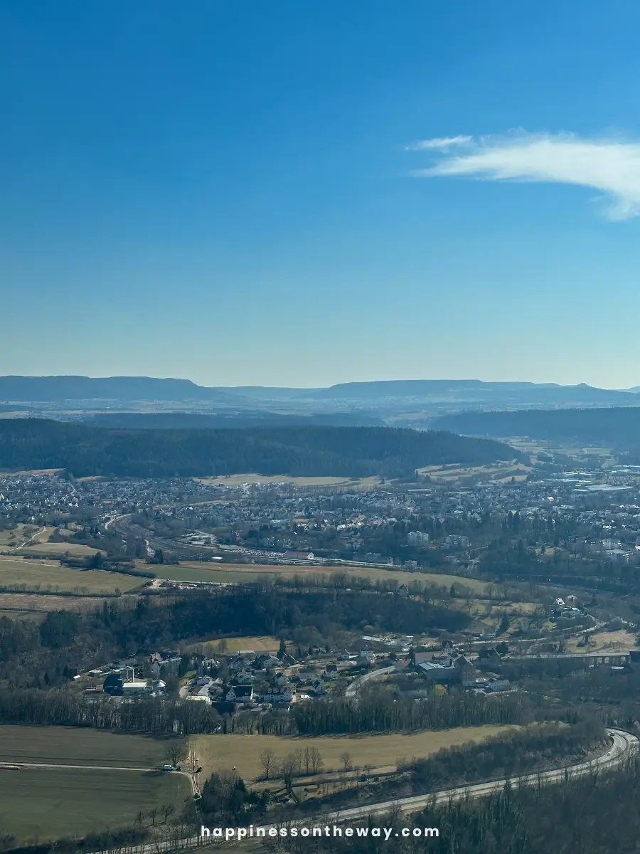 A panoramic view of Rottweil, Germany, from a high vantage point. Rolling hills, clusters of buildings, winding roads, and distant mountains stretch under a clear blue sky.