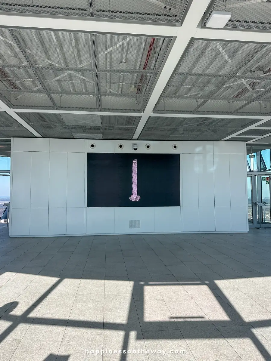 A modern, open indoor space with a large digital screen displaying a pink object showing the Rottweil Test Tower.