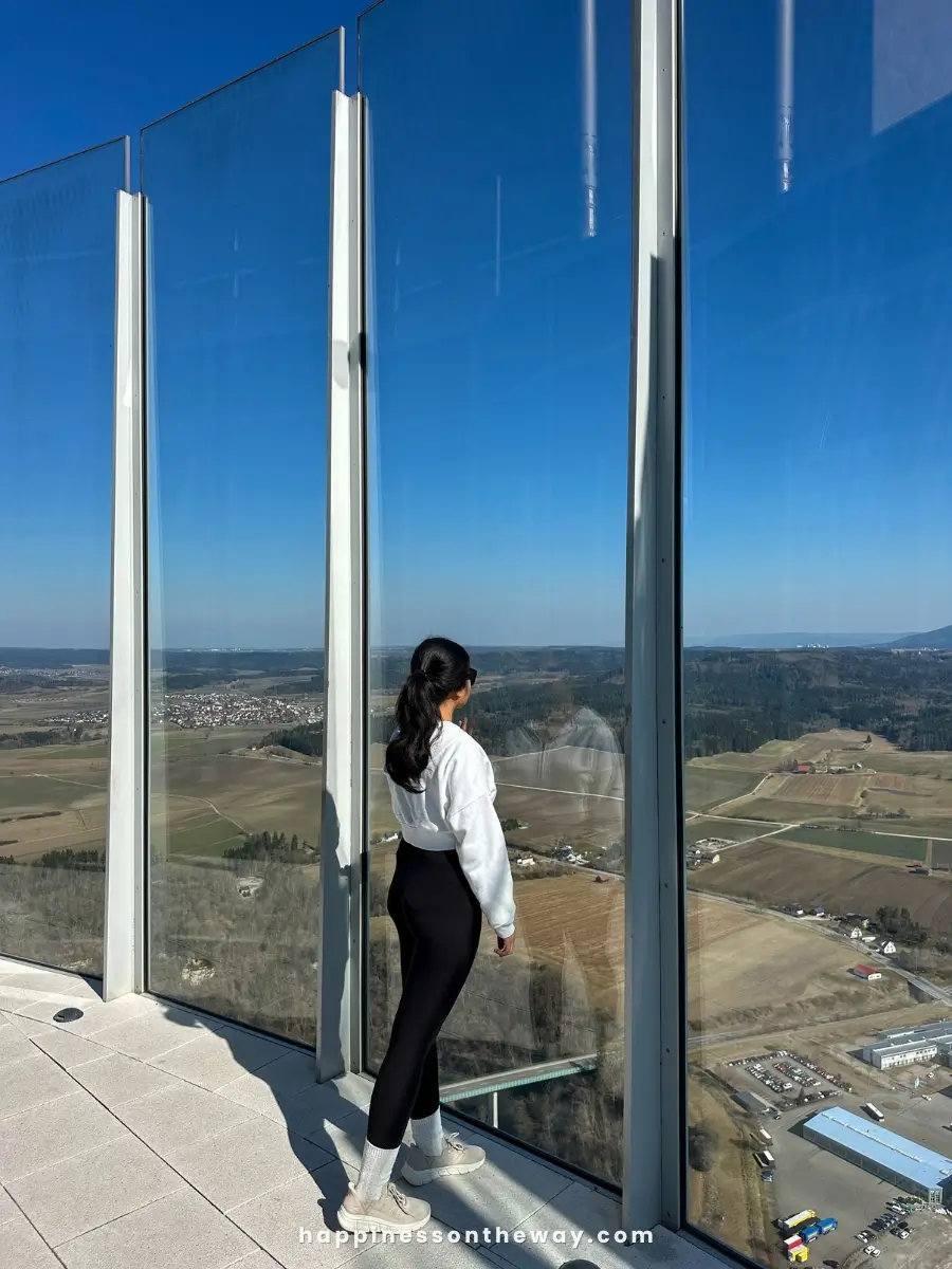 Me in a white jacket, black leggings, and white sneakers stands at a glass observation deck, gazing at the vast countryside and small towns below.