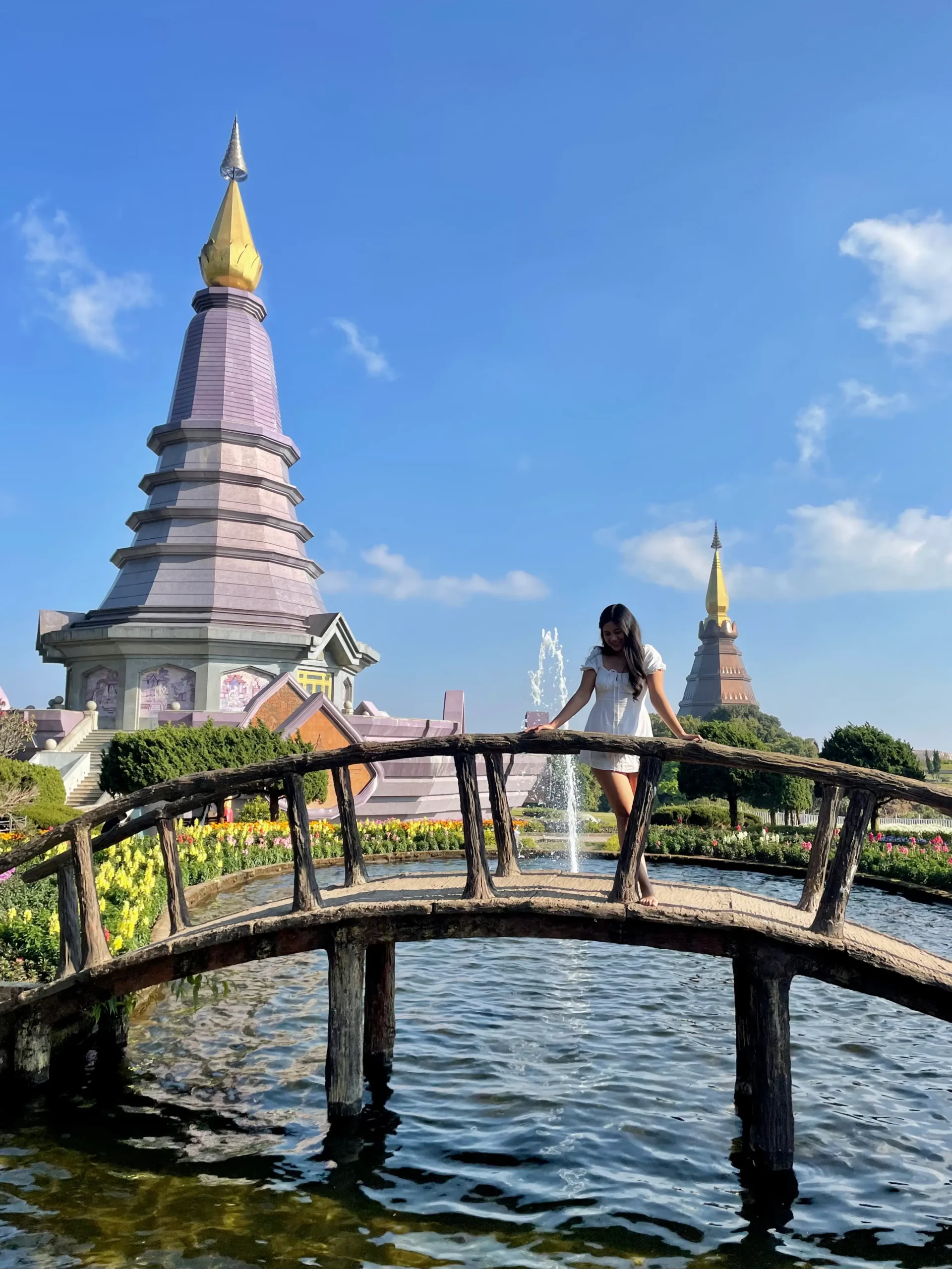 A woman in a white dress stands on a small arched wooden bridge over a pond, with two ornately designed pagodas and a garden full of flowers in Chiang Mai, Thailand.