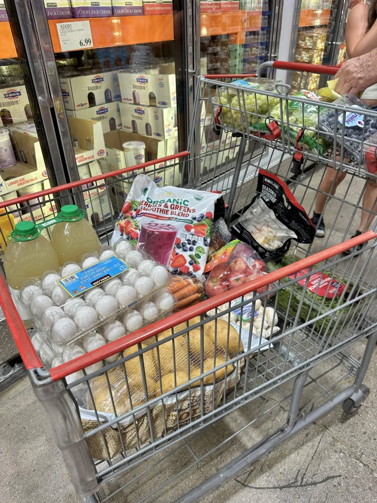 A shopping cart at Costco filled with groceries including eggs, lemonade, carrots, potatoes, smoothie blend, grapes, mushrooms, and other fresh produce.