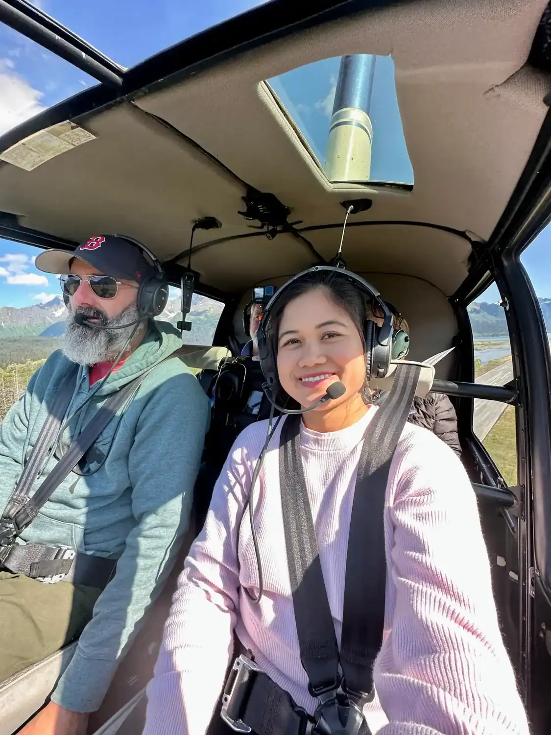 Gladis smiling inside a helicopter during a scenic ride in Alaska, wearing a headset and harness — part of a work exchange adventure while learning how to travel the world for free.