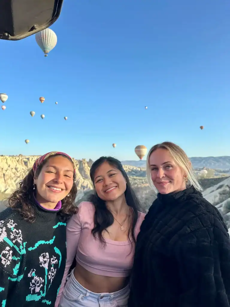 My covolunteers and I a stunning landscape dotted with dozens of colorful hot air balloons floating in the sky. The central figure wears a light pink crop top and white jeans, with scenic rocky formations of Cappadocia in the background.