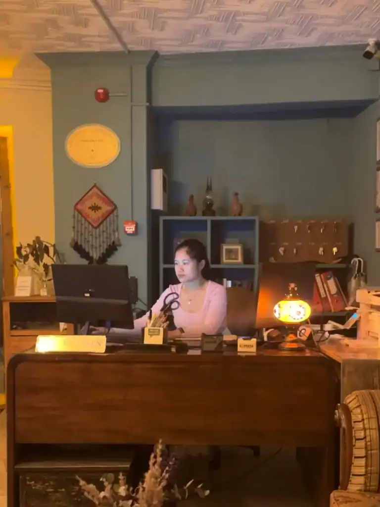 Day-to-day tasks while volunteering with Worldpackers. Sitting behind a wooden reception desk, focused on her computer in a warmly lit, vintage-style office. Behind her, shelves, key cubbies, and decorative pieces like a Turkish wall hanging and antique vases add a cozy, traditional vibe.