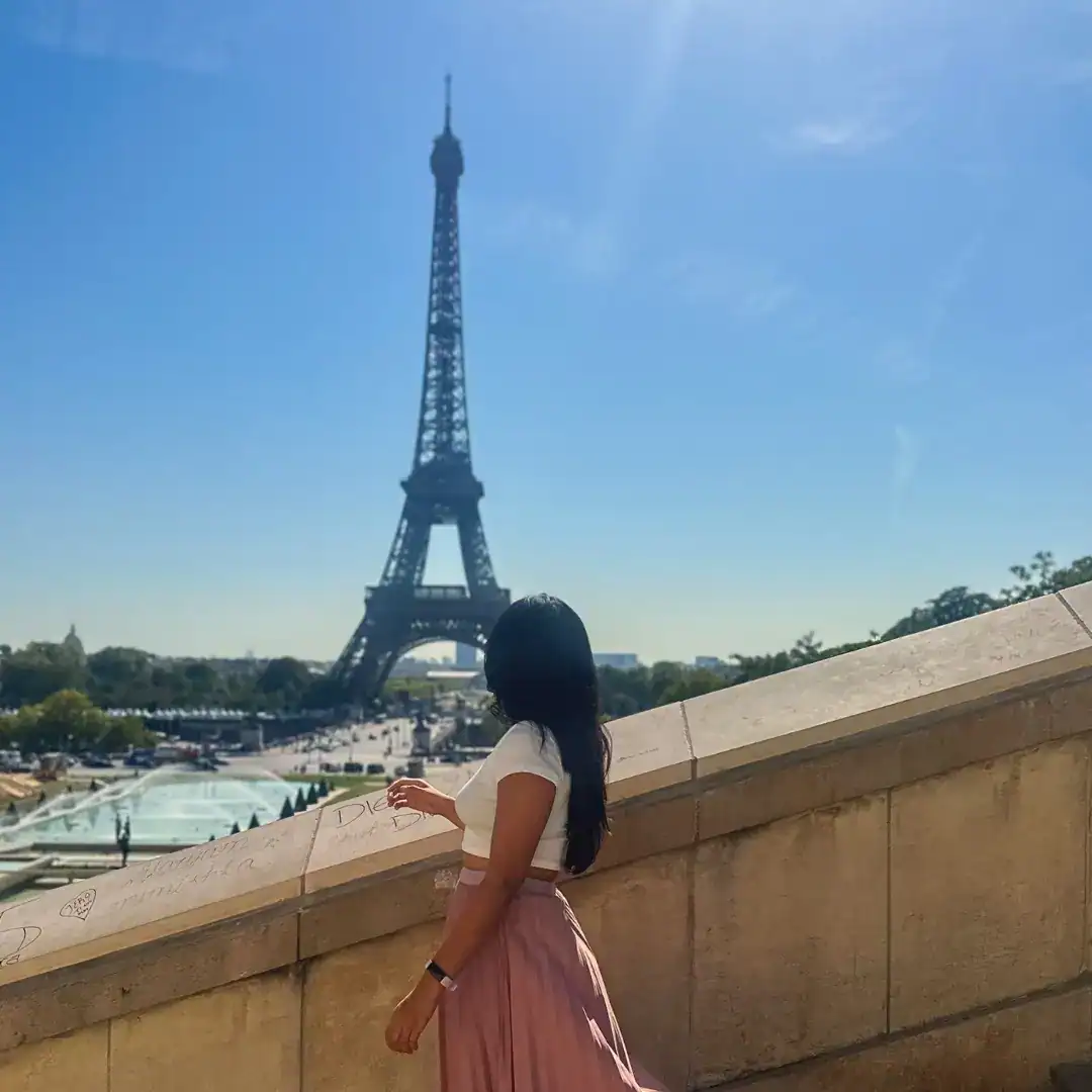 A woman with long dark hair, wearing a white crop top and flowing pink skirt, stands on a stone terrace gazing at the Eiffel Tower under a clear blue sky, showing the benefits of slow travel.