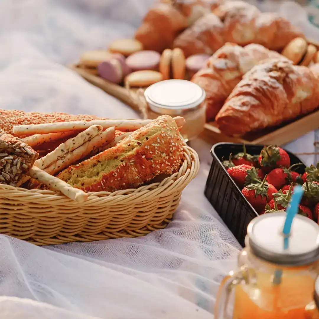 A cozy picnic spread on a soft white blanket features a basket of seeded breadsticks and rolls, a tray of croissants and macarons, fresh strawberries, a jar of honey, and a mason jar of orange juice with a blue straw. The warm lighting suggests a relaxed outdoor setting, possibly in Paris.