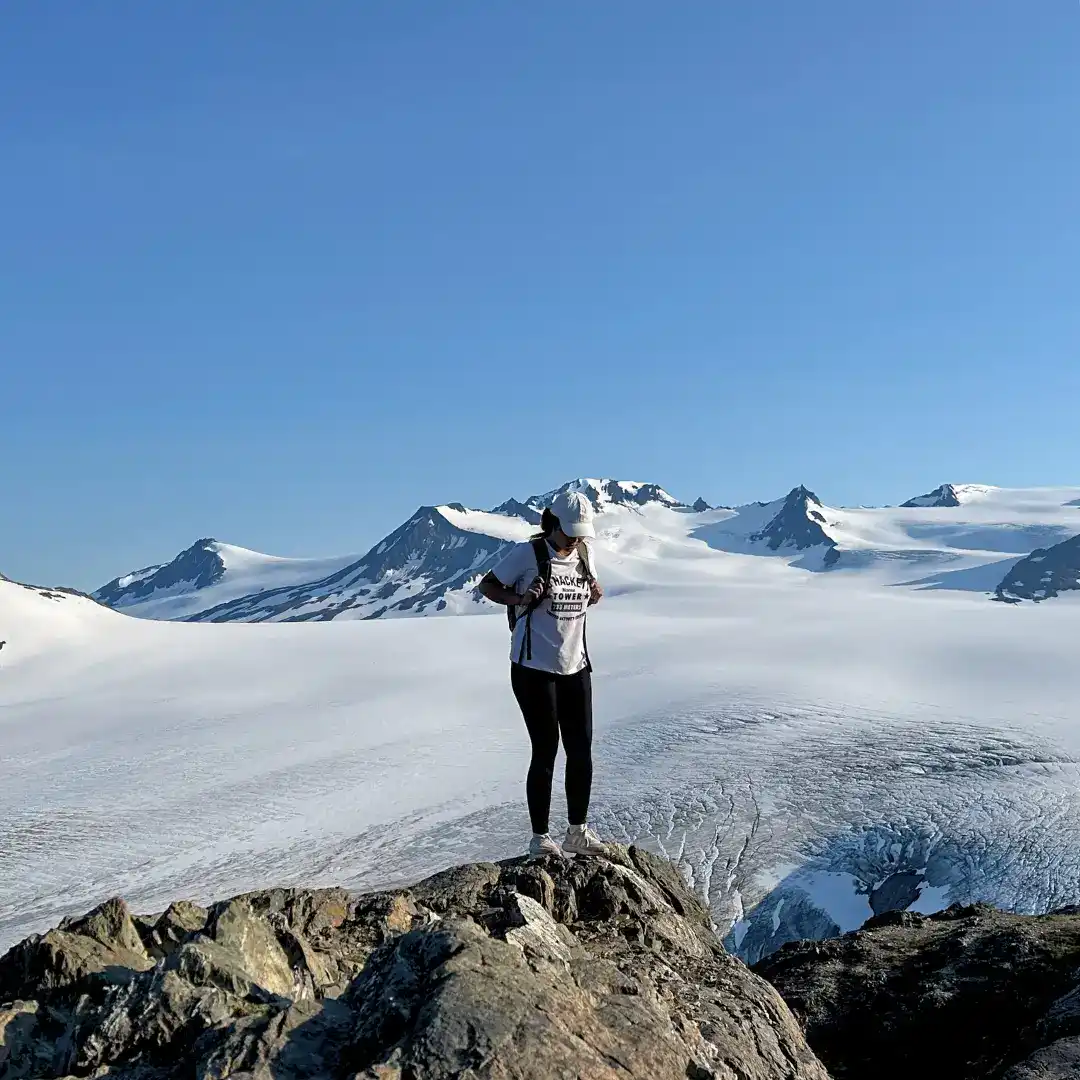A hiker stands atop a rocky outcrop, looking down at their gear, with a vast glacier and snow-covered mountain range stretching into the distance under a clear blue sky.
