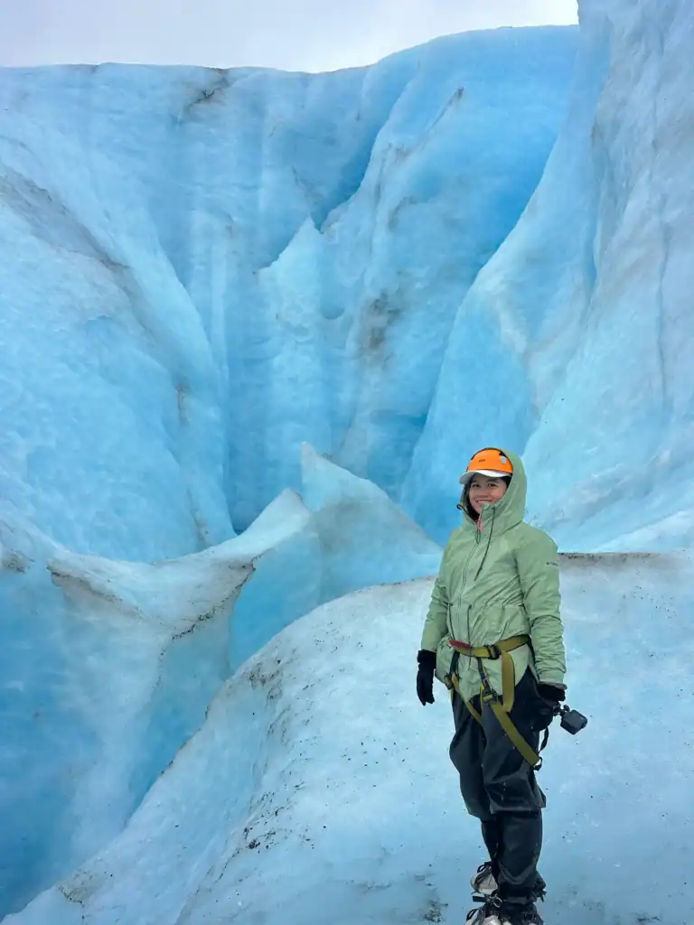 A person wearing a green jacket, crampons, and a helmet smiles while standing on a strikingly blue glacier, surrounded by deep crevasses and icy ridges. They appear prepared for ice hiking, holding a small camera and secured with a climbing harness.