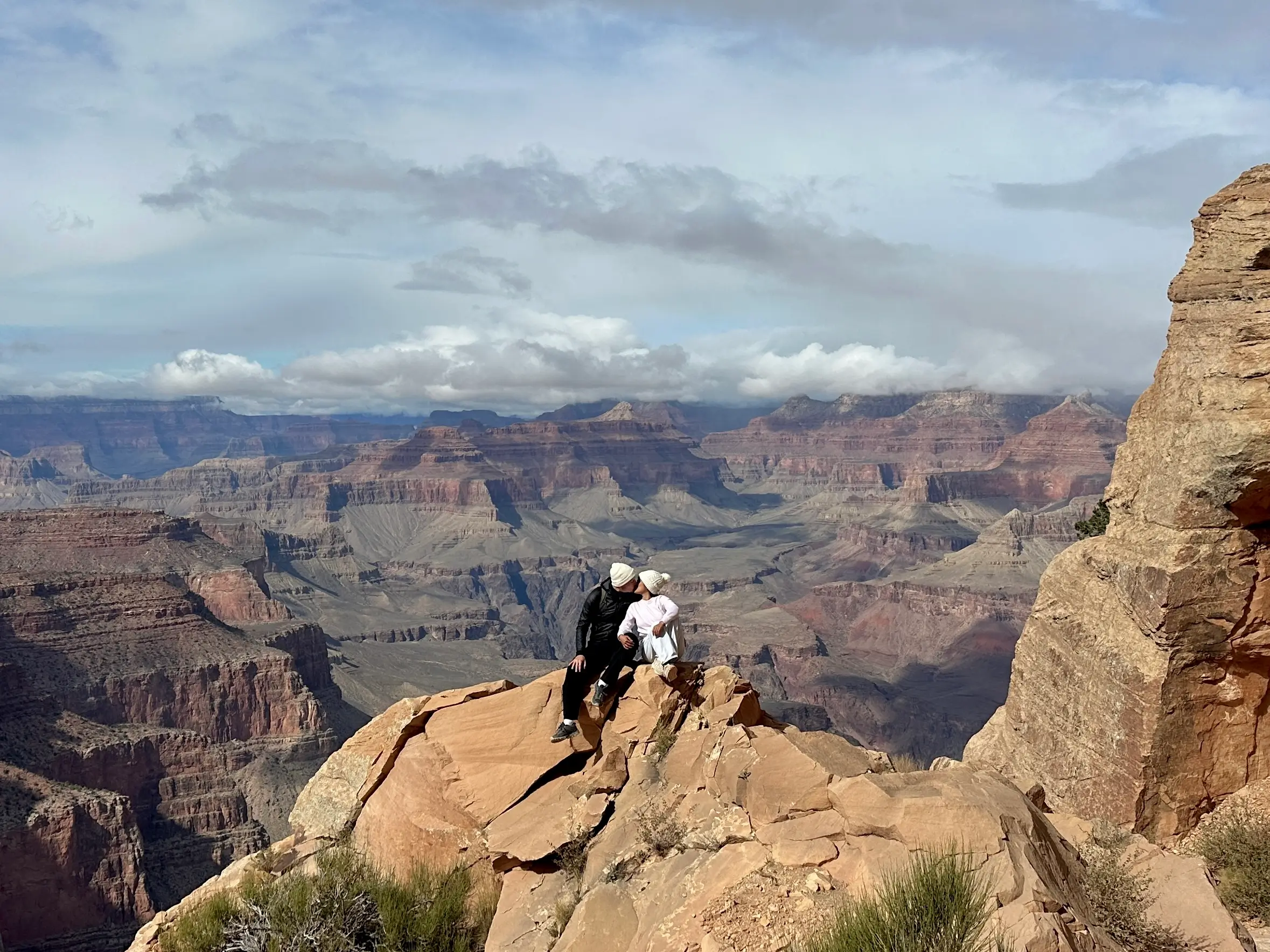 A couple sits close together on a rocky outcrop at the Grand Canyon, surrounded by sweeping canyon views under a partly cloudy sky.