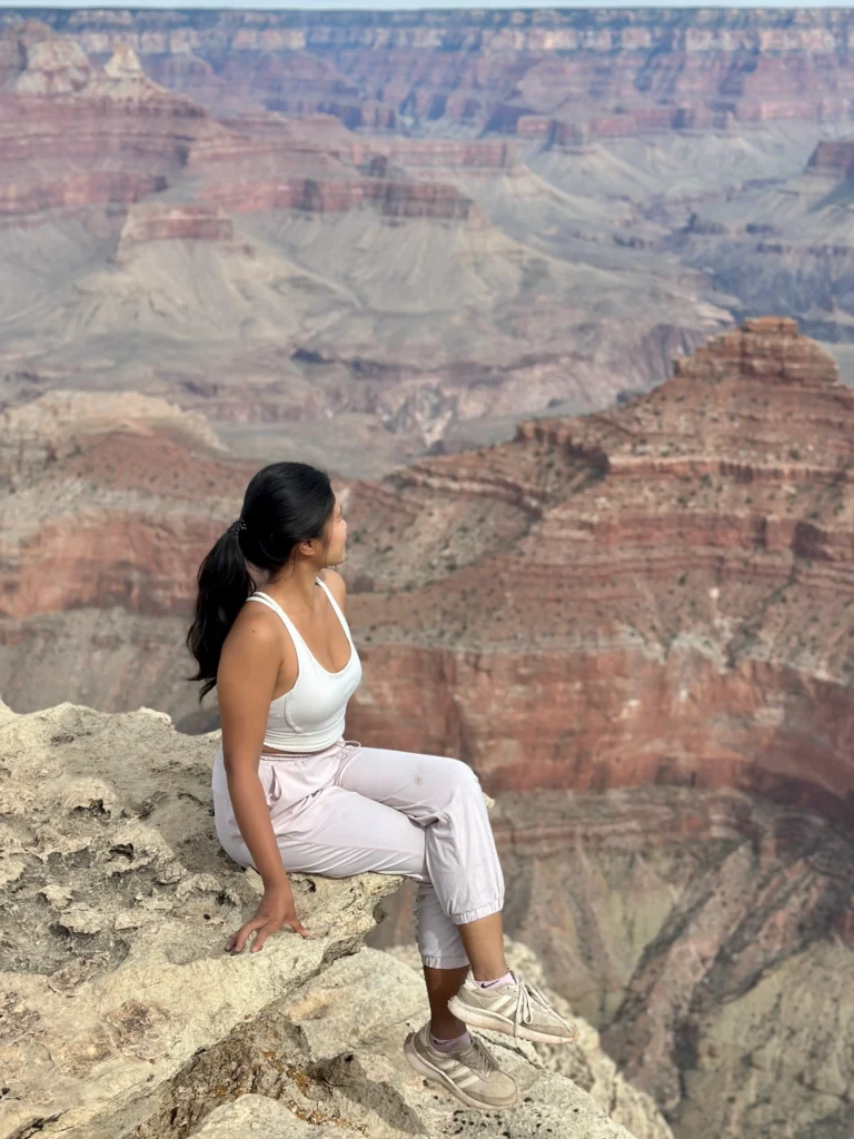 Me in light athletic wear sits on the edge of a rocky cliff, gazing out over the layered red and tan rock formations of the Grand Canyon.