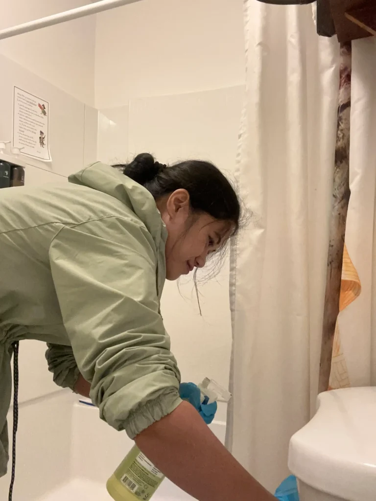 A woman in a light green jacket and blue gloves cleaning a bathroom toilet with a spray bottle, leaning over near a shower curtain. A printed sign with cleaning instructions is visible on the tiled wall