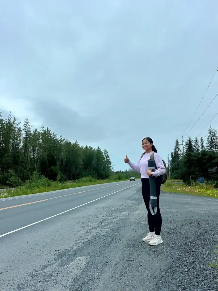 Gladis standing by the roadside in Alaska with her thumb out, hitchhiking while solo traveling—part of her journey learning how to travel the world for free