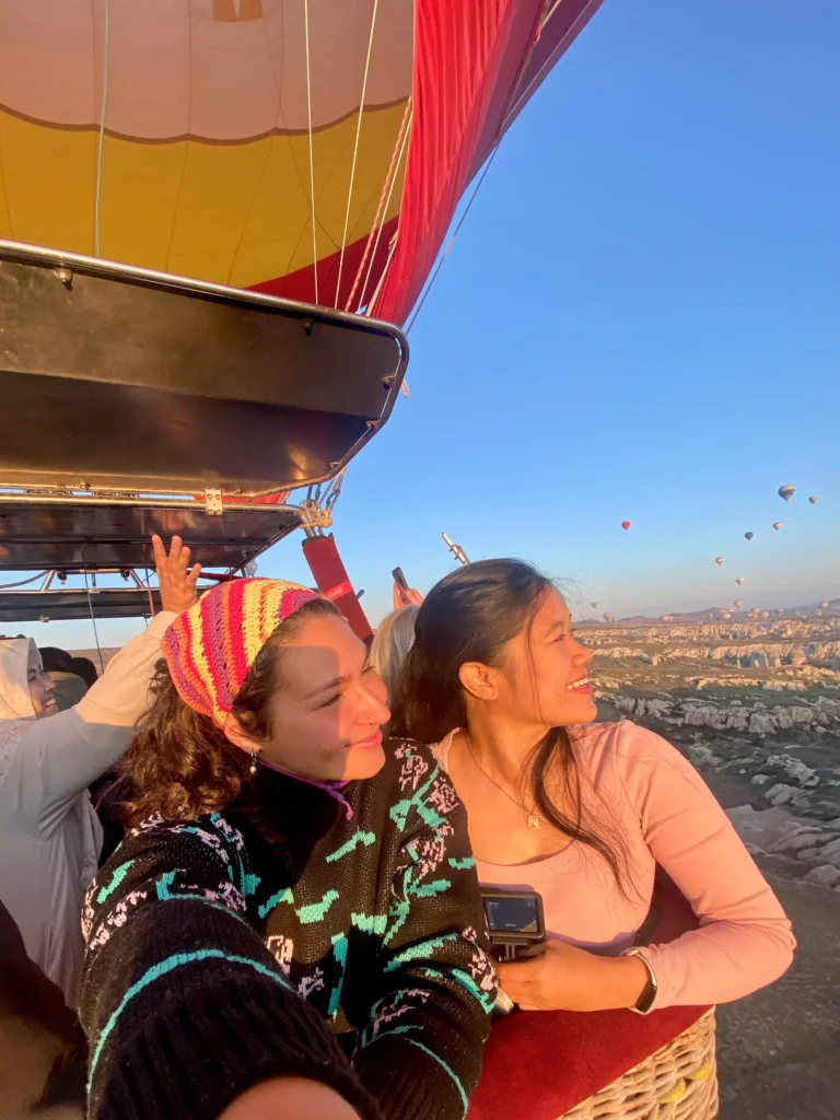 My covolunteer and I smiling and looking out from a hot air balloon basket during sunrise in Cappadocia, with other balloons floating in the background over a rocky landscape.