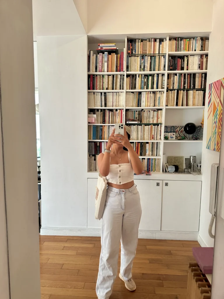 I'm taking a mirror selfie in a stylish room with wood floors and tall bookshelves, wearing a white crop top and loose white pants, holding a tote bag.