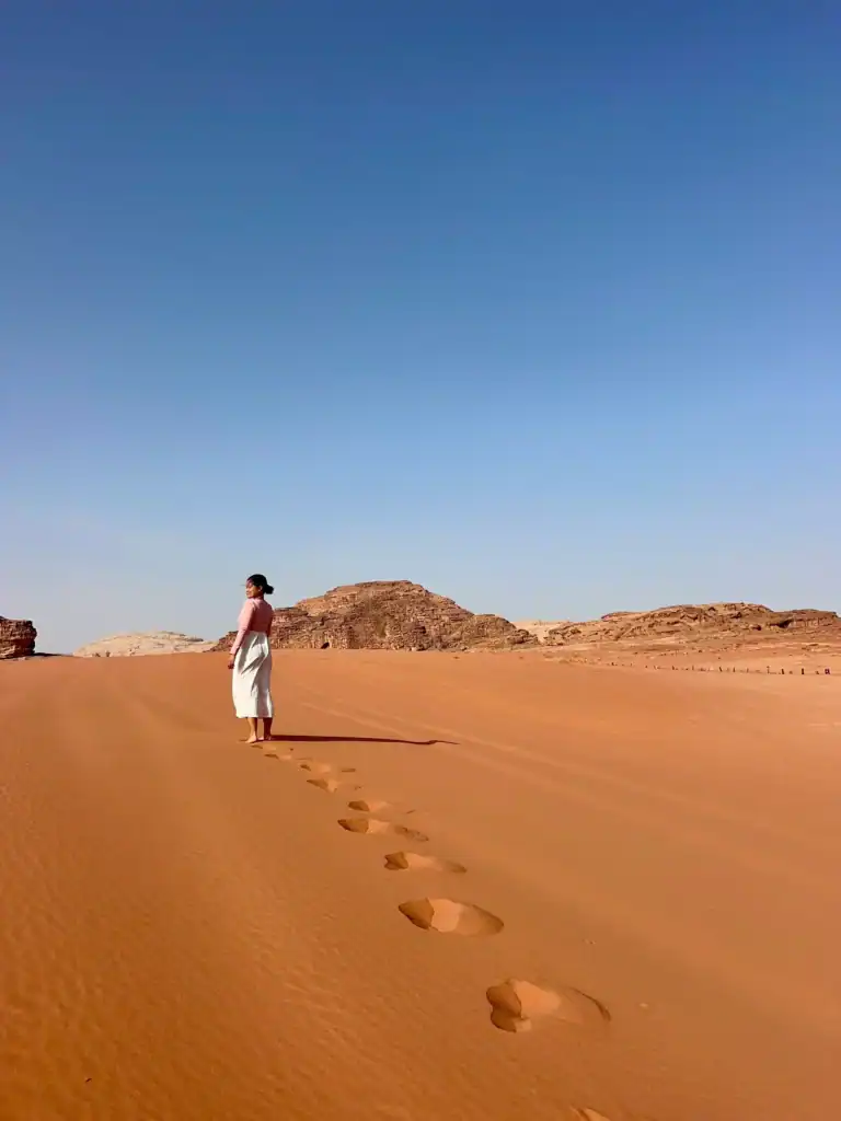 Woman walking barefoot on red sand dunes in Wadi Rum, Jordan, leaving a trail of footprints under a clear blue sky.