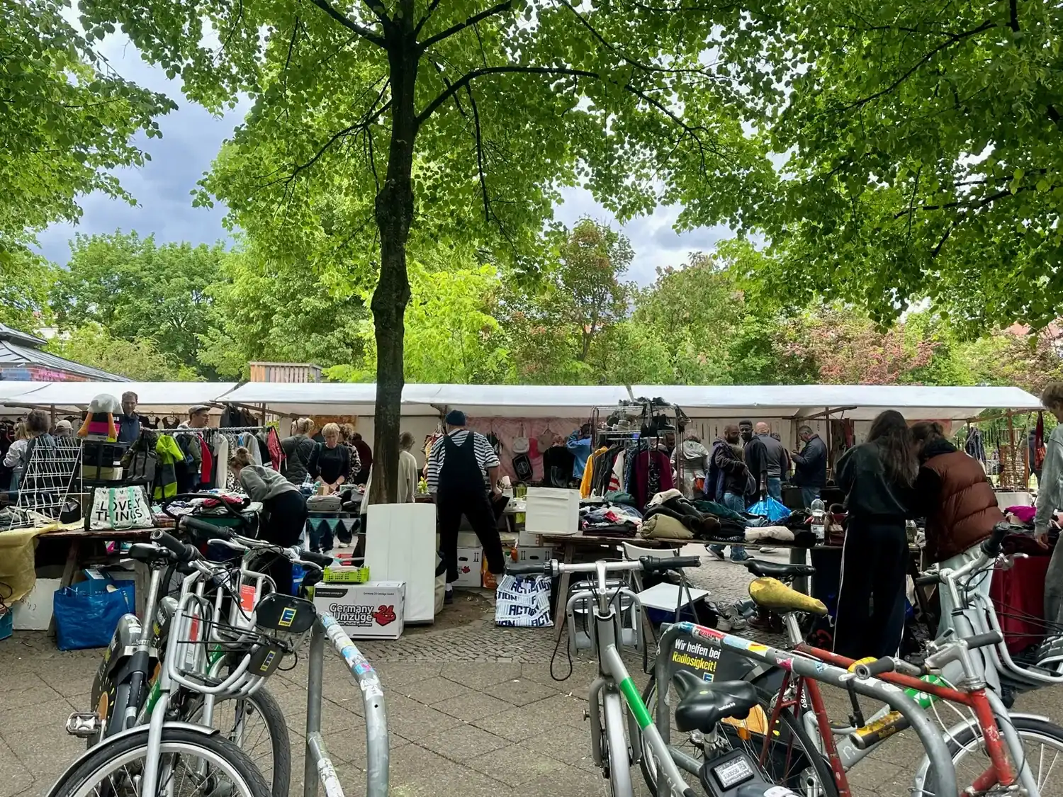 Outdoor flea market under leafy trees in Berlin, with people browsing and bicycles parked nearby.