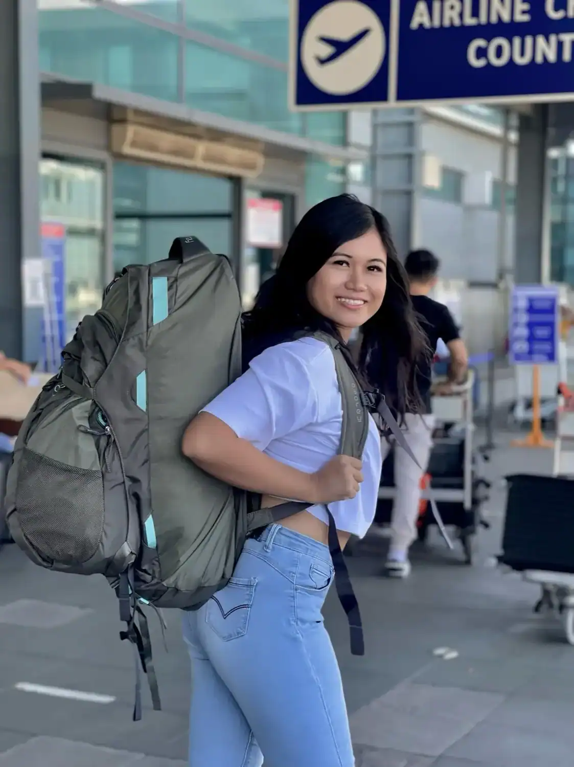 Woman with a large backpack smiling outside an airport, symbolizing how to choose a travel destination with excitement and readiness to explore.