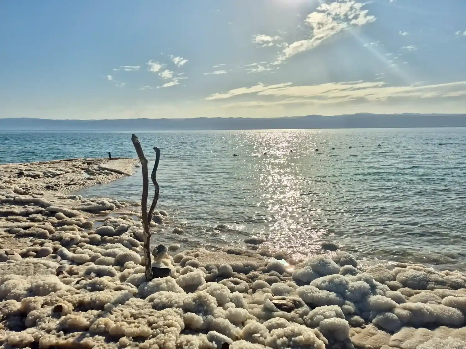 Crystalline salt formations line the shore of the Dead Sea under a bright afternoon sun, with calm turquoise waters.