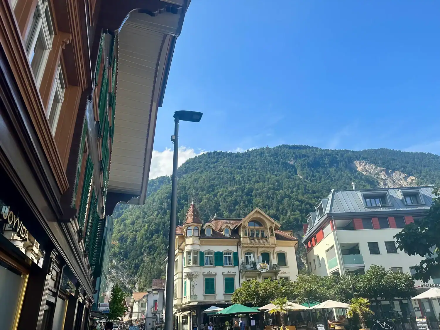 Street view in Interlaken, Switzerland, showing charming European buildings with alpine architecture set against a backdrop of green forested mountains.