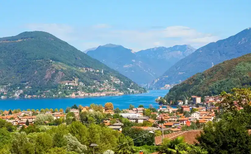 Scenic view of Lake Lugano surrounded by lush green mountains and dotted with charming towns featuring terracotta rooftops.