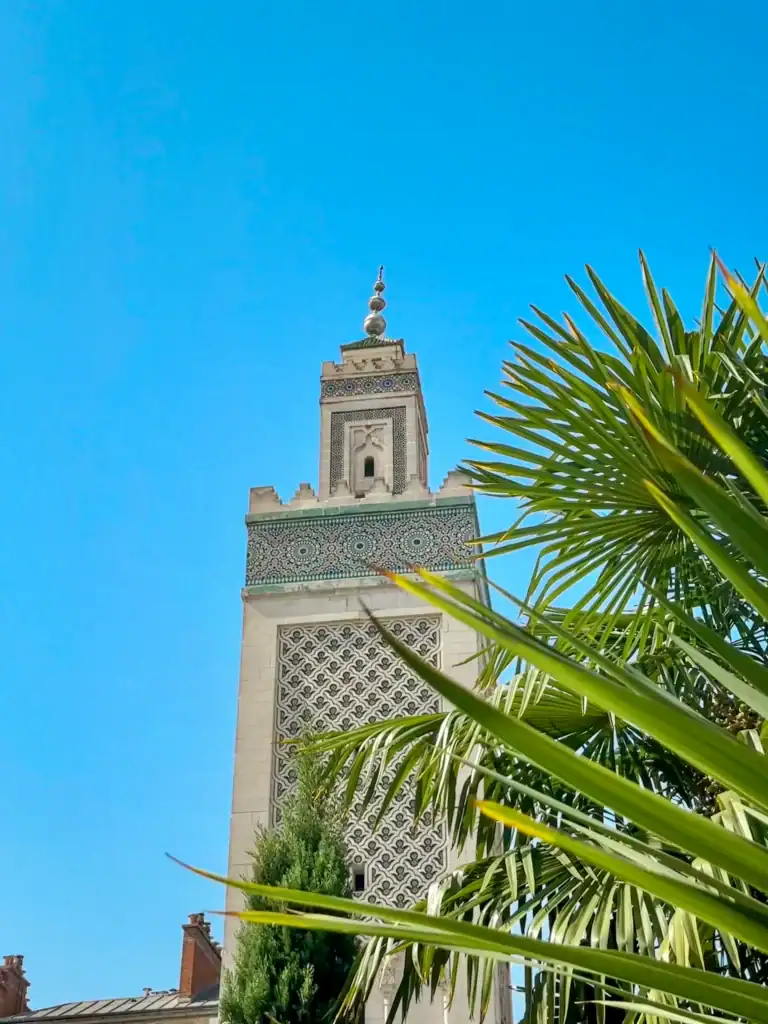 A tall, intricately decorated minaret of the Grand Mosque of Paris rises against a vivid blue sky, partially framed by lush palm fronds and greenery.