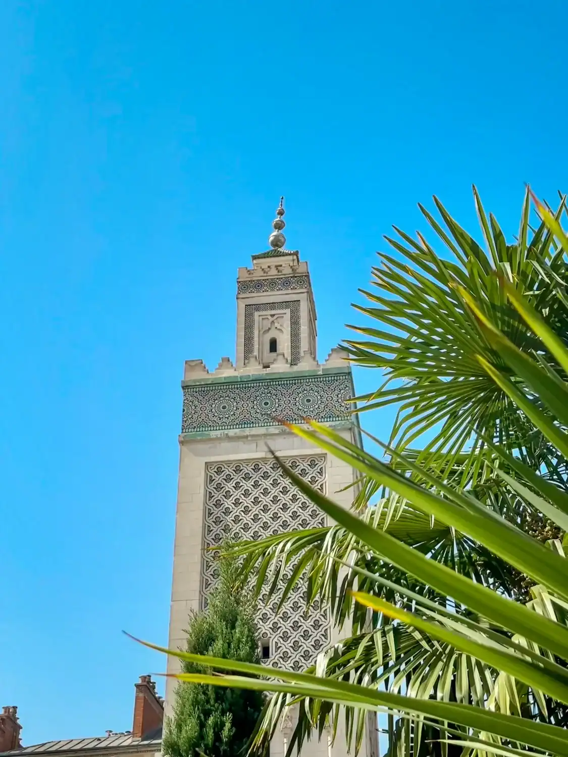 A tall, intricately decorated minaret of the Grand Mosque of Paris rises against a vivid blue sky, partially framed by lush palm fronds and greenery.