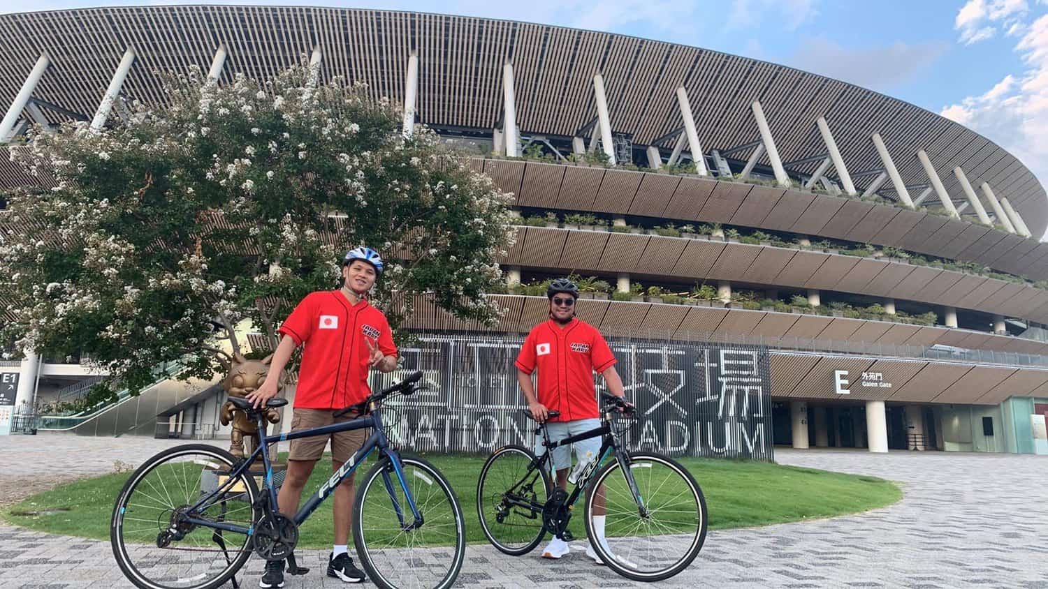 My brother and my friend in red jerseys with bikes in front of Japan National Stadium during a guided tour in Tokyo.