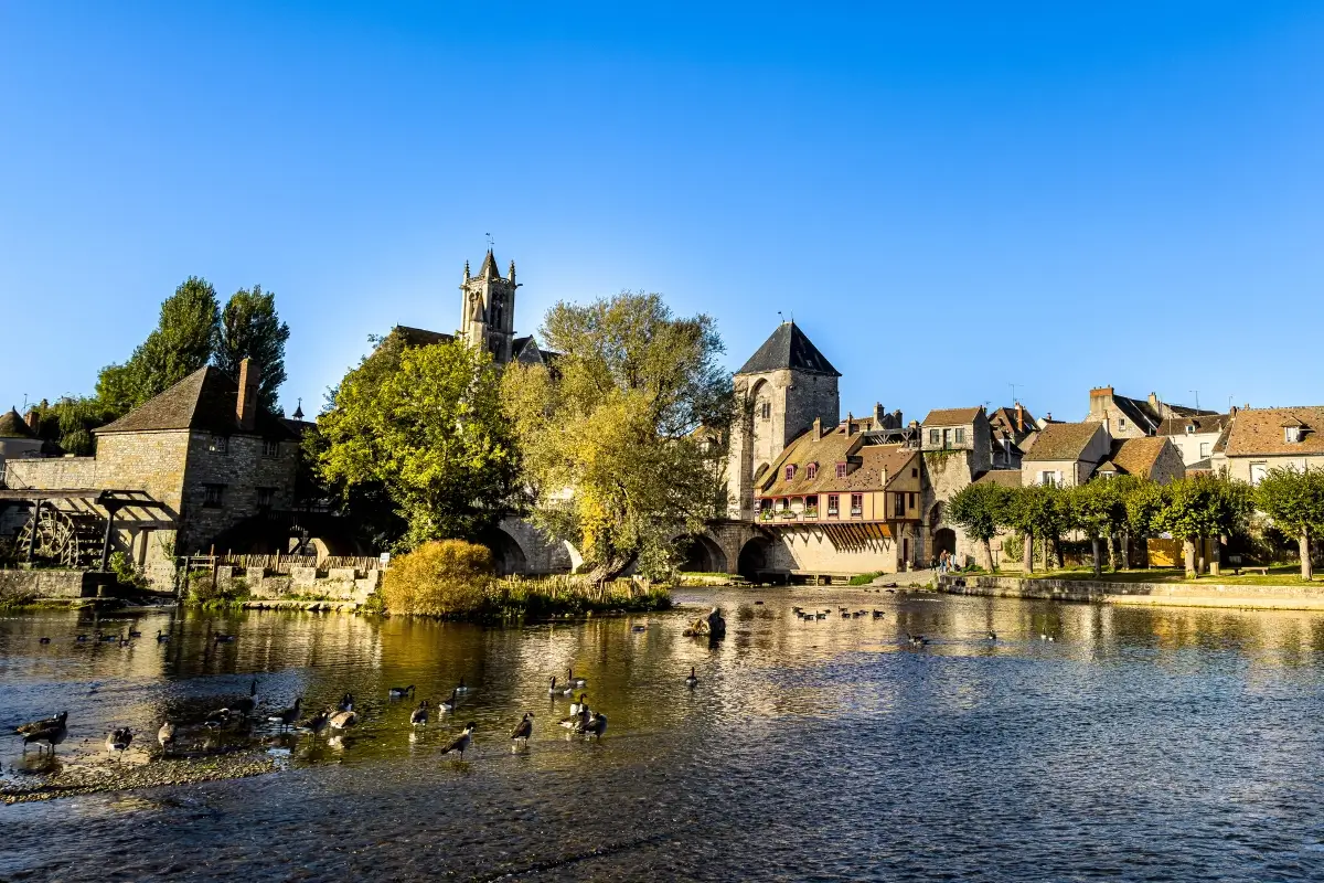 Scenic riverside view of Moret-sur-Loing with historic stone buildings, ducks in the water, and a medieval bridge in the background