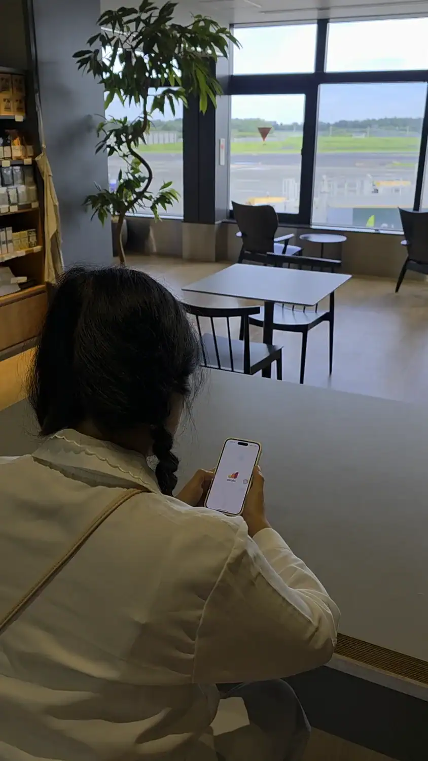 Woman at an airport café browses her phone with an eSIM app open, likely selecting the best eSIM for Japan before her flight.
