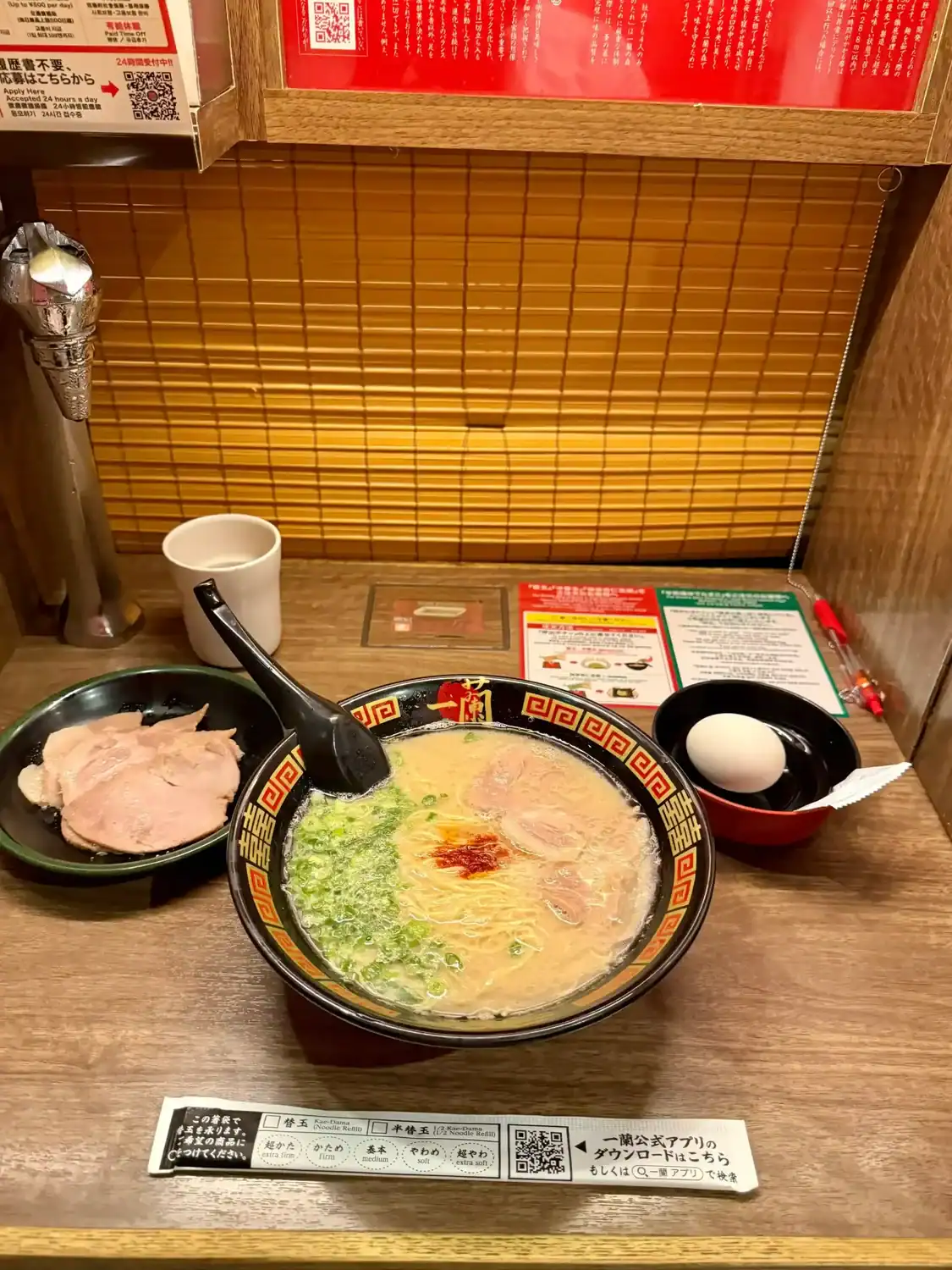 A steaming bowl of tonkotsu ramen is served in a private booth at Ichiran, featuring thin noodles in a rich pork bone broth, topped with sliced pork, chopped green onions, and a dollop of red chili paste.
