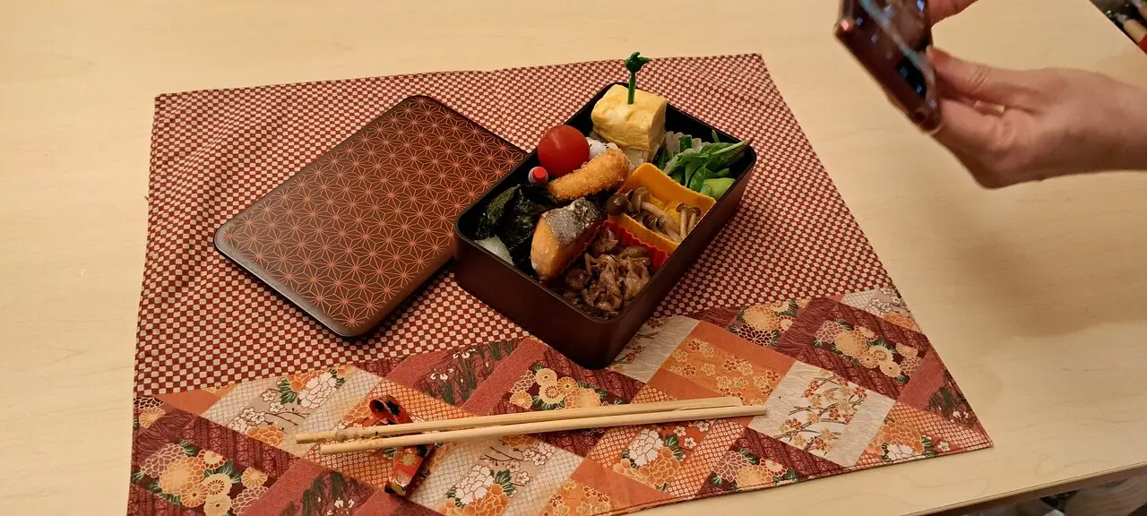 A finished bento box filled with fish, vegetables, egg, and side dishes on a patterned placemat, with someone taking a photo of it.