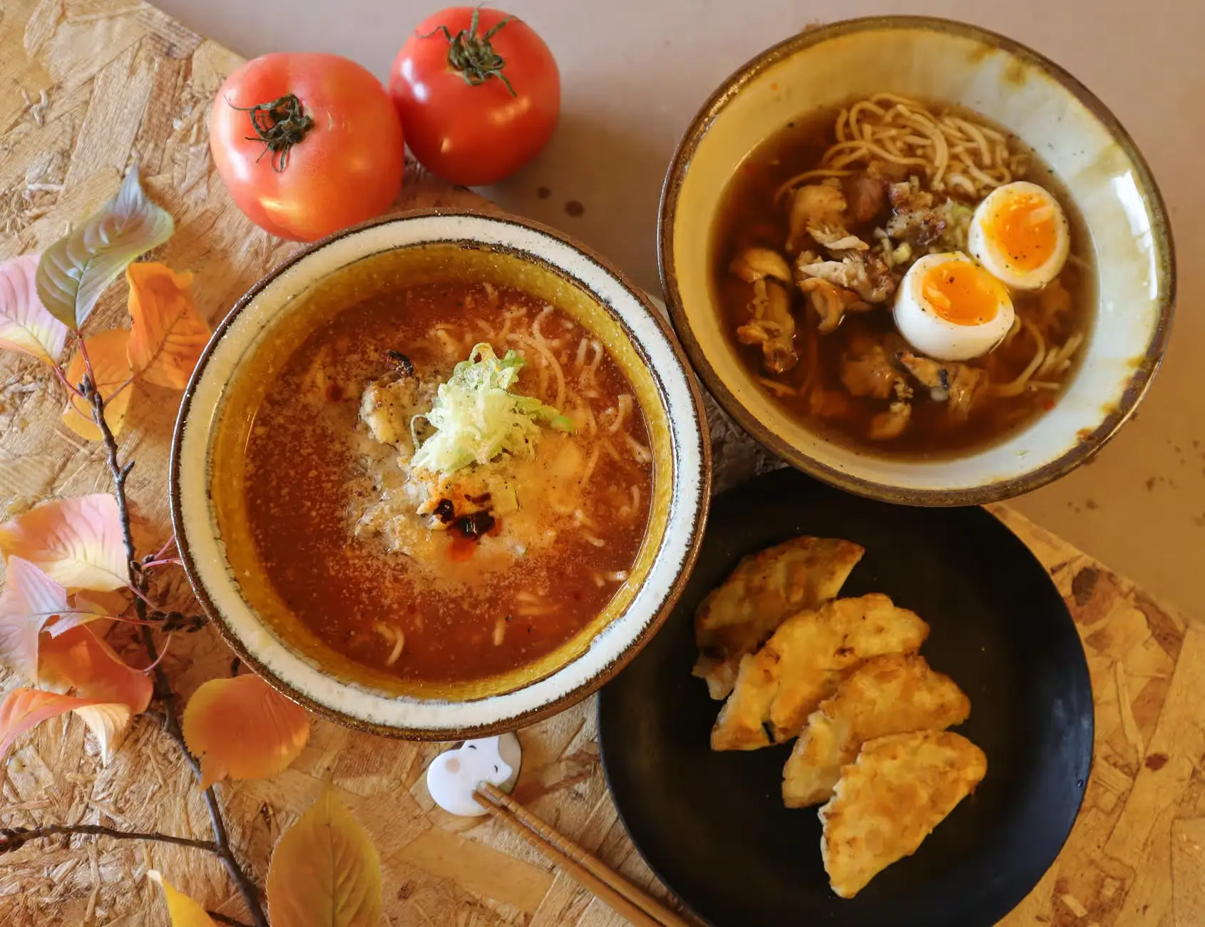 Two bowls of ramen, one spicy and one clear broth, served with a plate of pan-fried gyoza on a wooden table with tomatoes and decorative autumn leaves. One of the best cooking classes in Osaka 