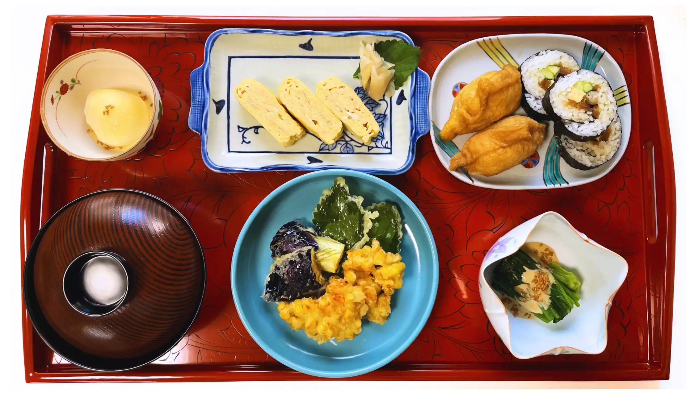 A traditional Japanese home-style meal served on a red tray, featuring tamagoyaki, inari sushi, rolled sushi, vegetable tempura, miso soup, and a spinach side dish.