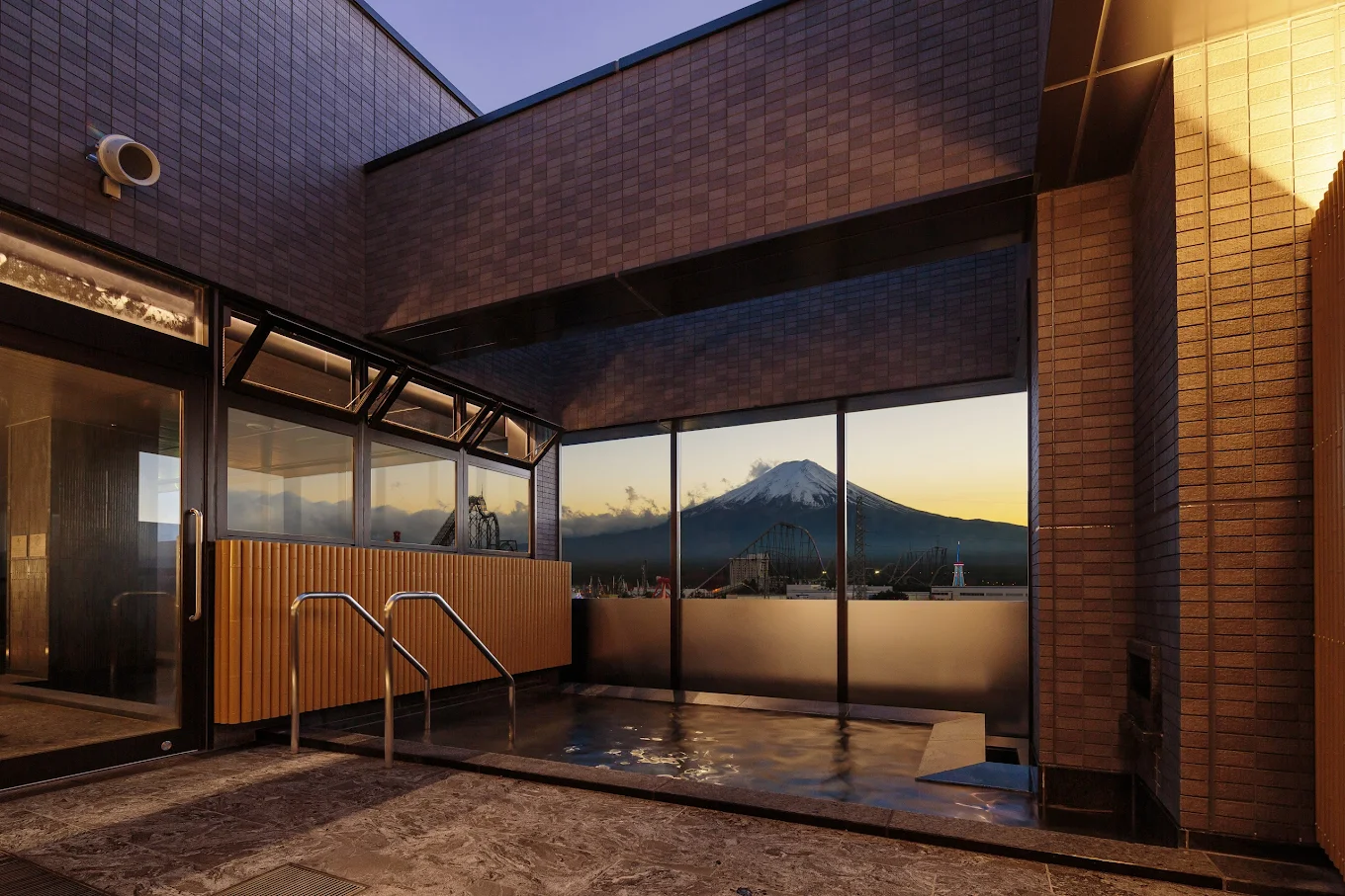 A serene rooftop onsen with warm lighting and modern tile walls overlooks a majestic snow-capped mountain at sunset.