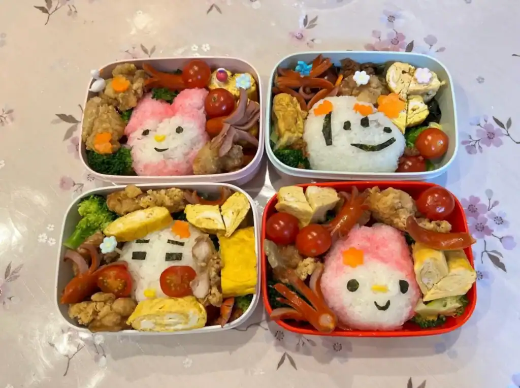Four colorful bento lunch boxes with character-shaped rice balls, cherry tomatoes, rolled omelet, fried chicken pieces, broccoli, and sausage arranged in a cute style.