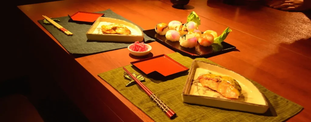 Izakaya-style Japanese dishes served on a wooden table, including grilled fish, temari sushi, and pickled ginger, arranged on traditional mats.