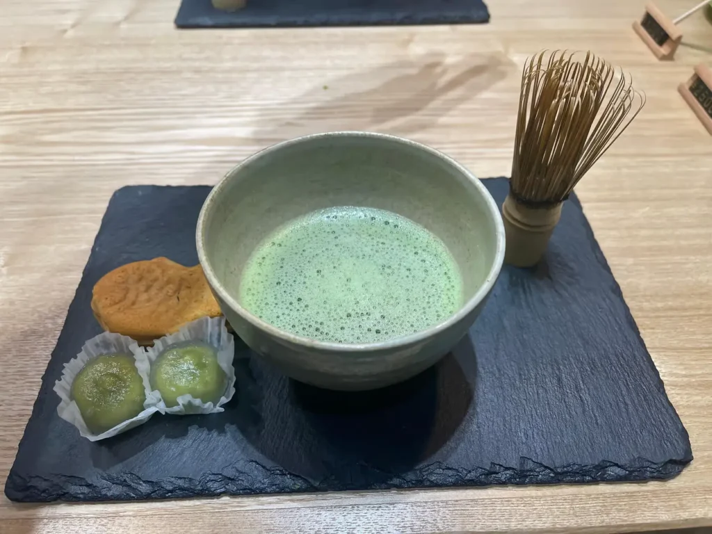 A bowl of freshly whisked matcha with a bamboo whisk, served alongside small Japanese sweets on a black slate plate.