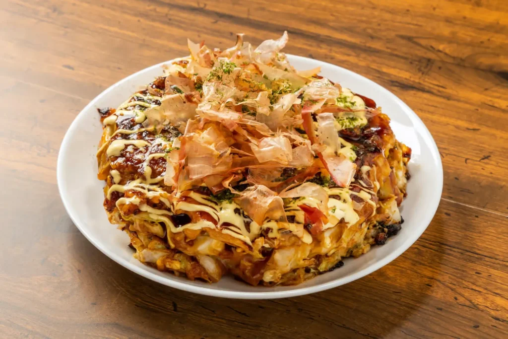 A freshly cooked okonomiyaki topped with bonito flakes, mayonnaise, and sauce, served on a white plate.