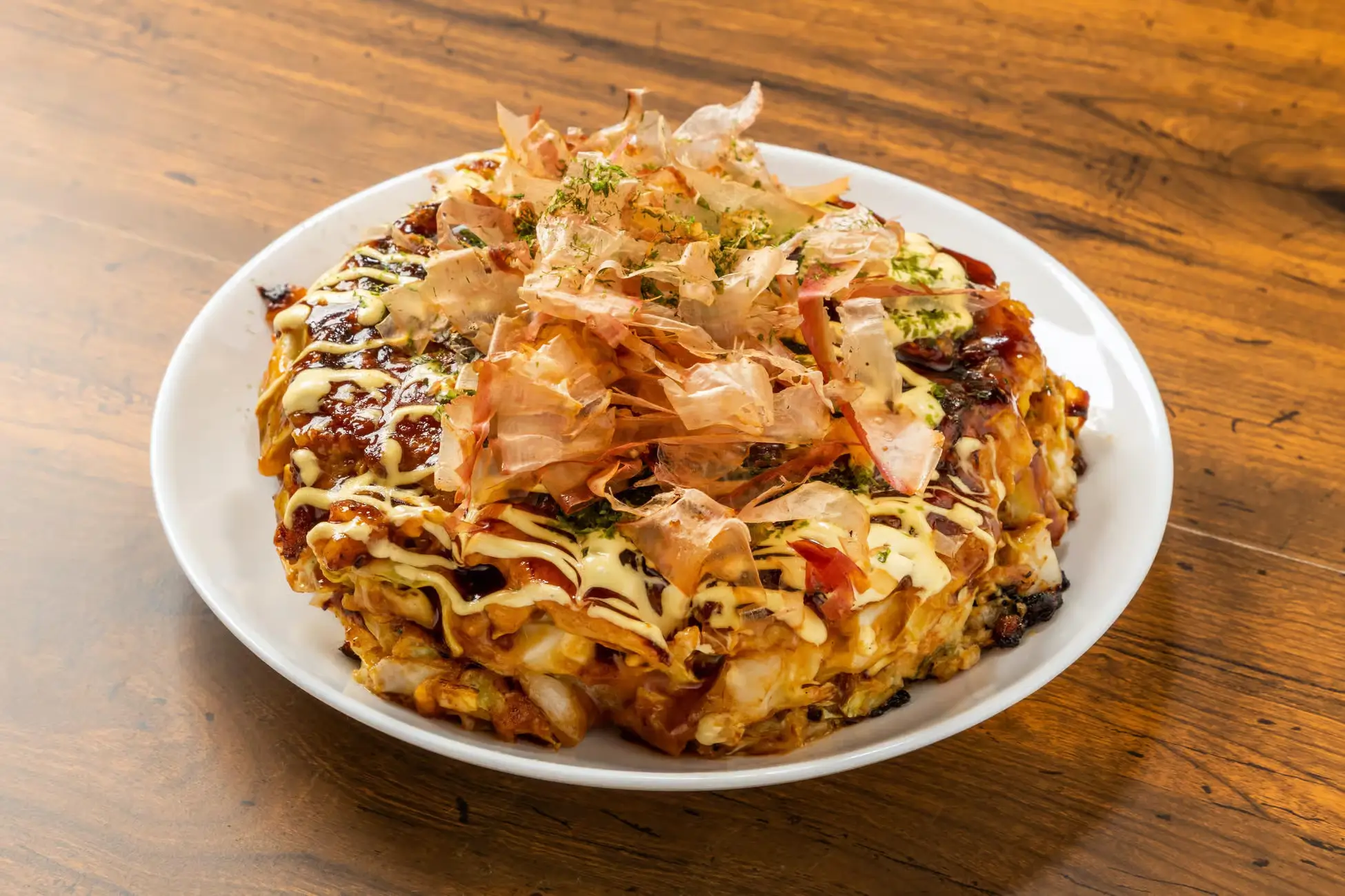 A freshly cooked okonomiyaki topped with bonito flakes, mayonnaise, and sauce, served on a white plate.