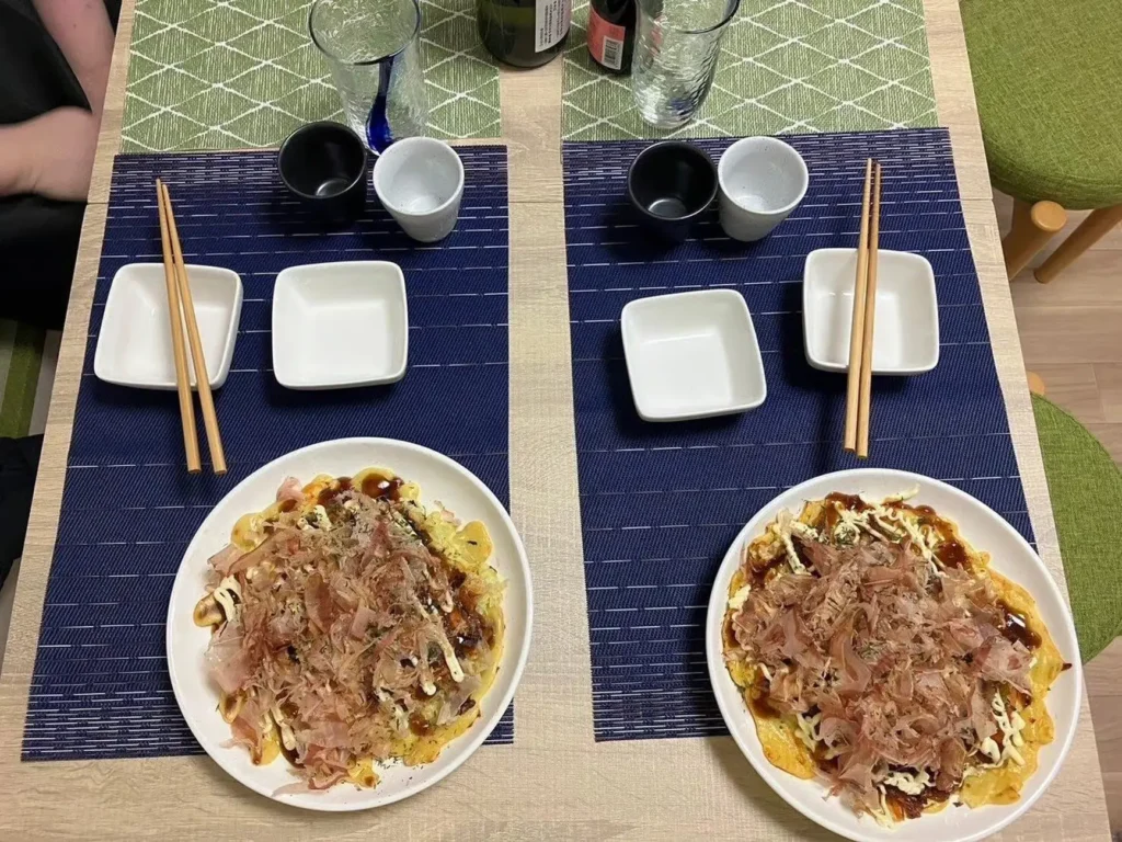 Two plates of okonomiyaki topped with bonito flakes set on a dining table with chopsticks, small dishes, and drinks prepared for tasting.