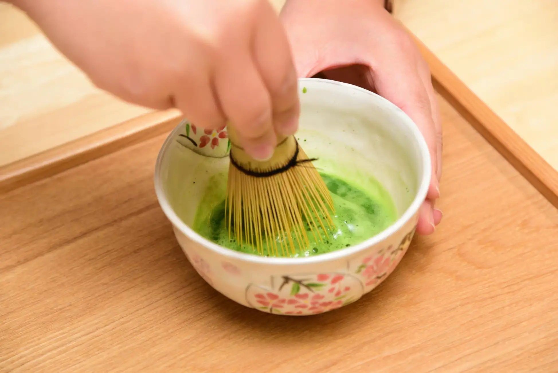 Hands whisking bright green matcha tea in a floral ceramic bowl during a tea ceremony.