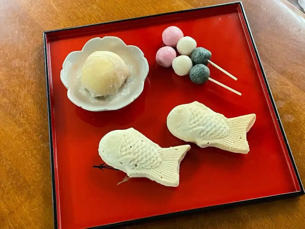 Traditional wagashi sweets on a red tray, including daifuku, taiyaki-shaped confections, and colorful dango on skewers.