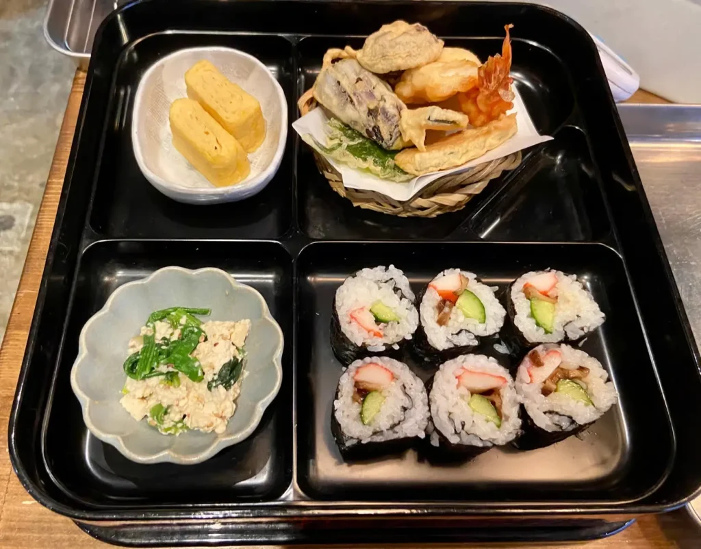 Bento box from a washoku cooking class, featuring rolled sushi, tamagoyaki, tempura, and a tofu side dish arranged neatly in separate compartments.