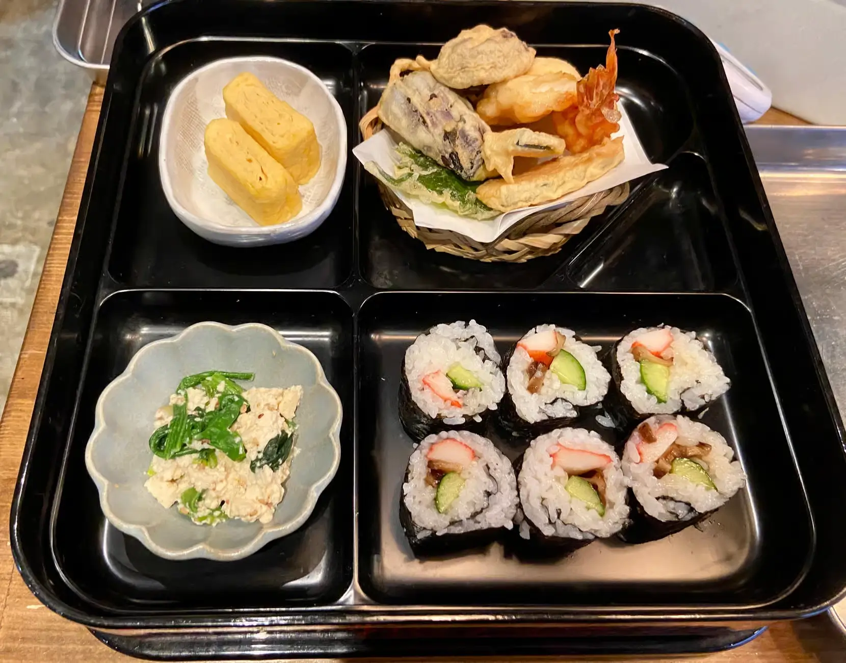 Bento box from a washoku cooking class, featuring rolled sushi, tamagoyaki, tempura, and a tofu side dish arranged neatly in separate compartments.