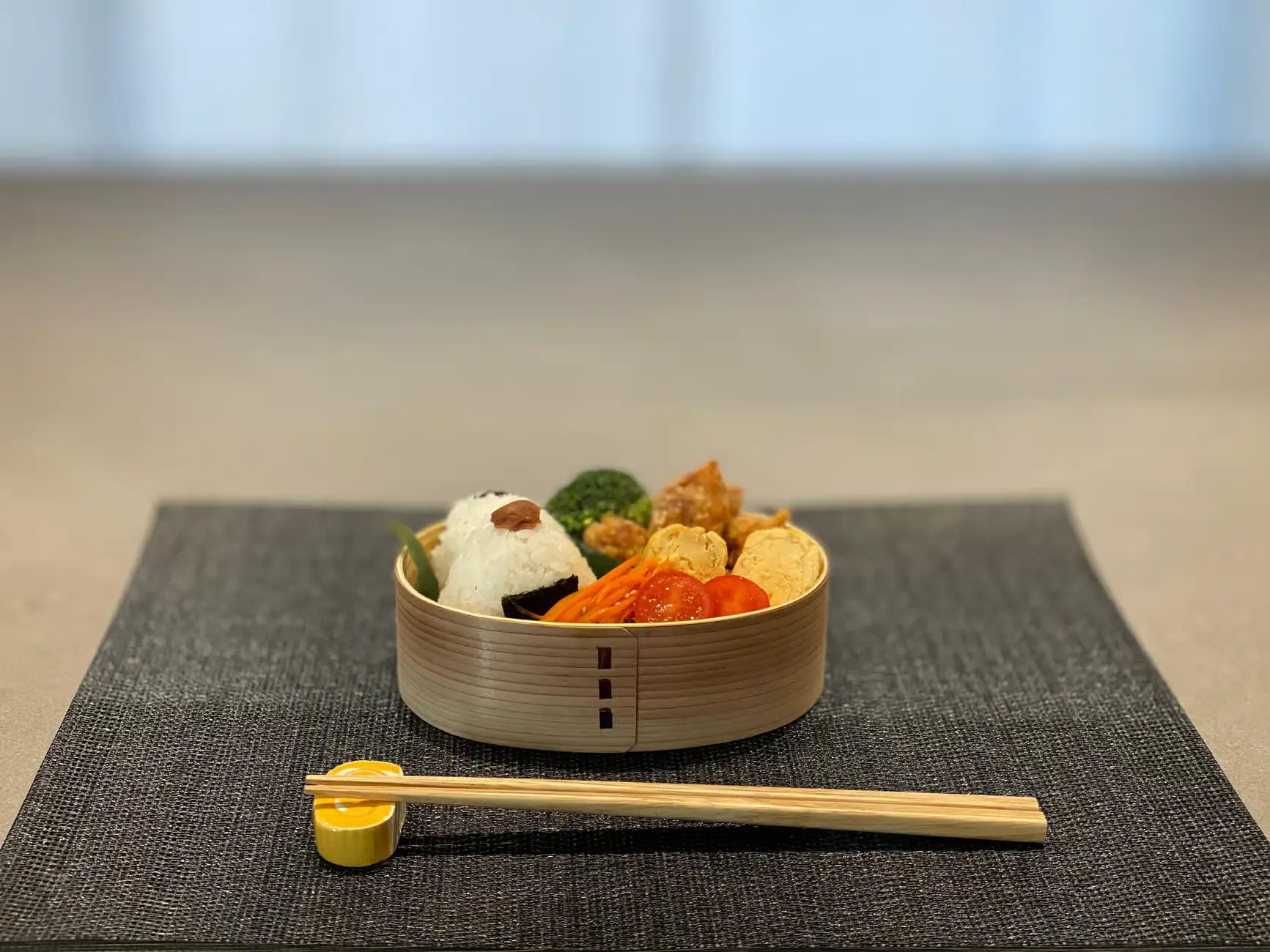 A traditional Japanese bento box filled with onigiri rice ball, tamagoyaki, cherry tomatoes, carrots, broccoli, and fried chicken.