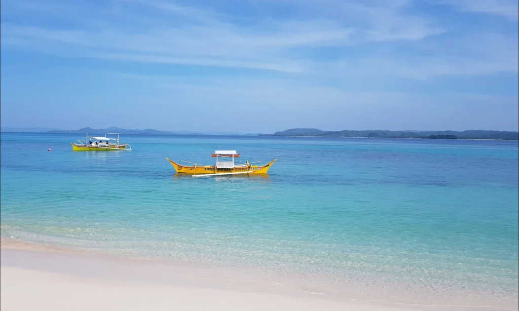 Two small yellow boats are floating on clear turquoise water near a white sandy shore under a bright blue sky at Daku Beach. One of the famous beach in Siargao