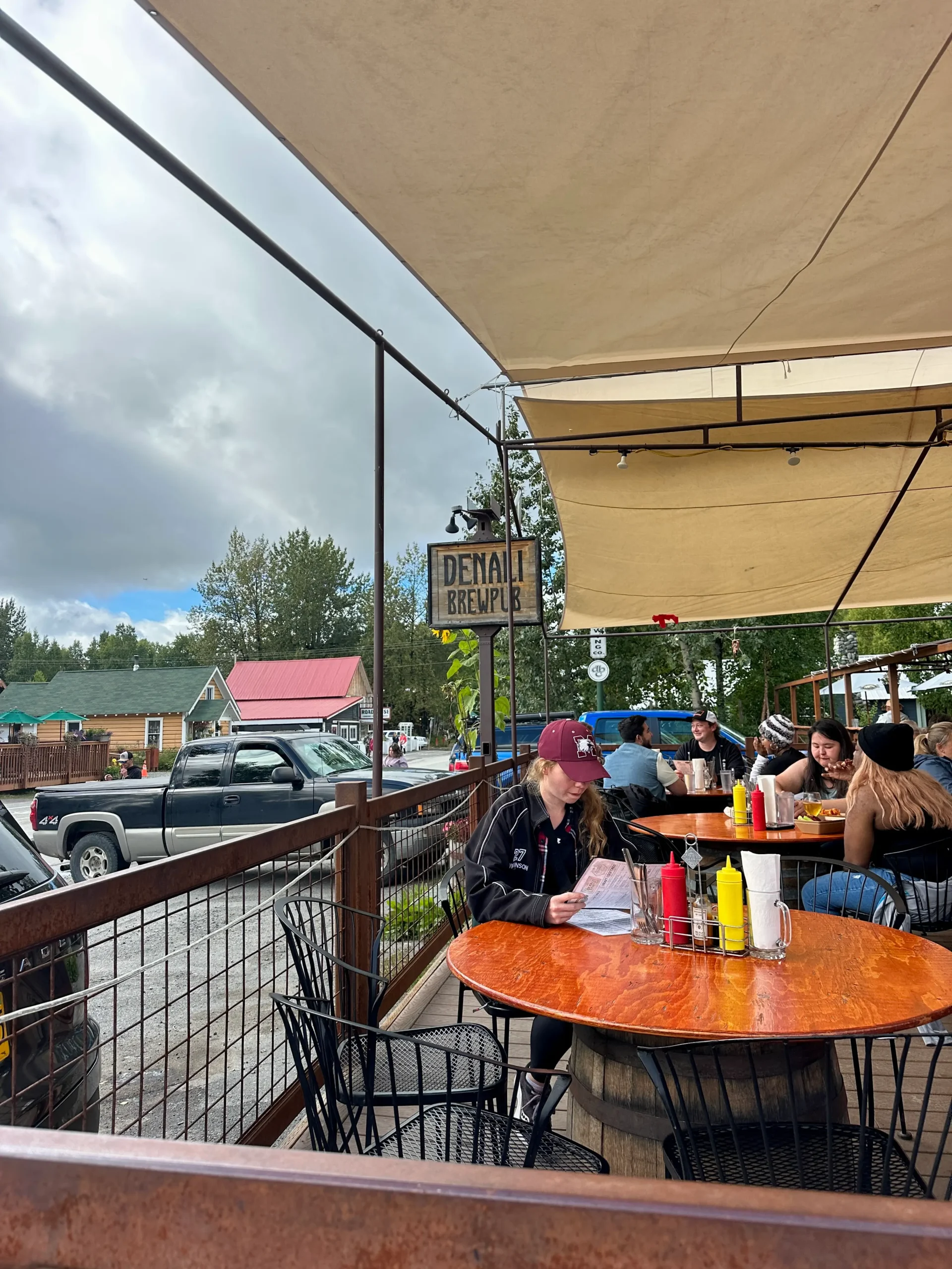 Outdoor patio at Denali Brewery showing a round wooden table with condiments and a woman in a red cap sitting alone under a shaded canopy.