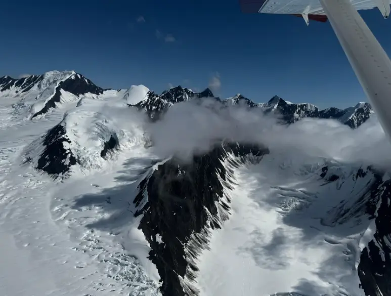 A small aircraft wing visible over the snow-covered mountain of Denali, Alaska, with clouds drifting between rugged peaks.