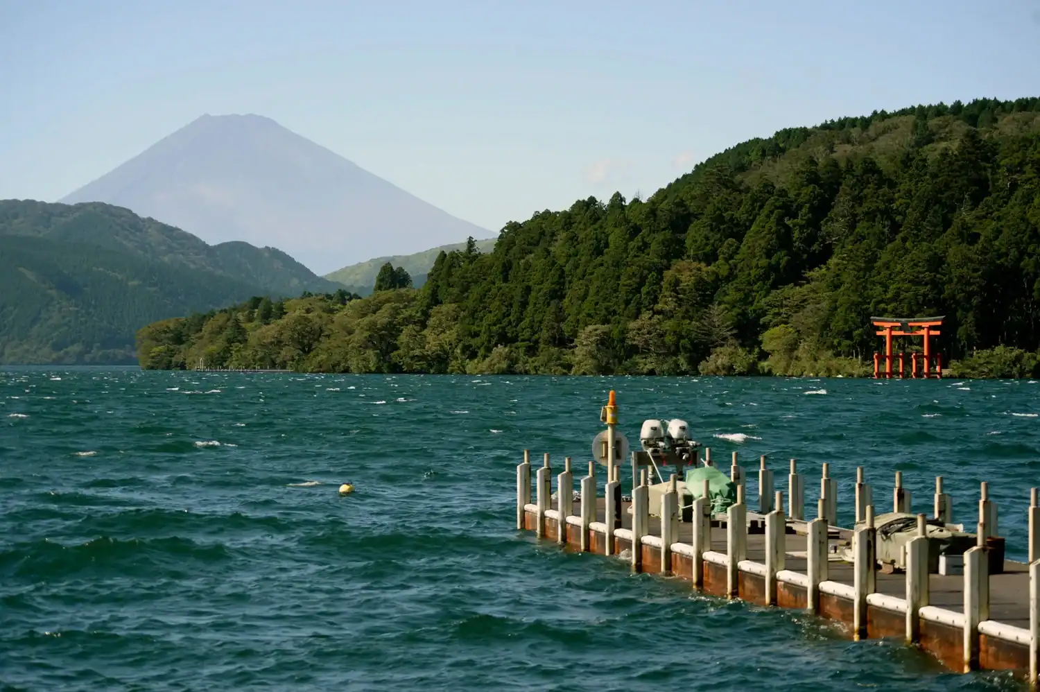 Lake Ashi in Hakone with Mount Fuji in the distance, a red torii gate visible across the water, forested mountains on both shores